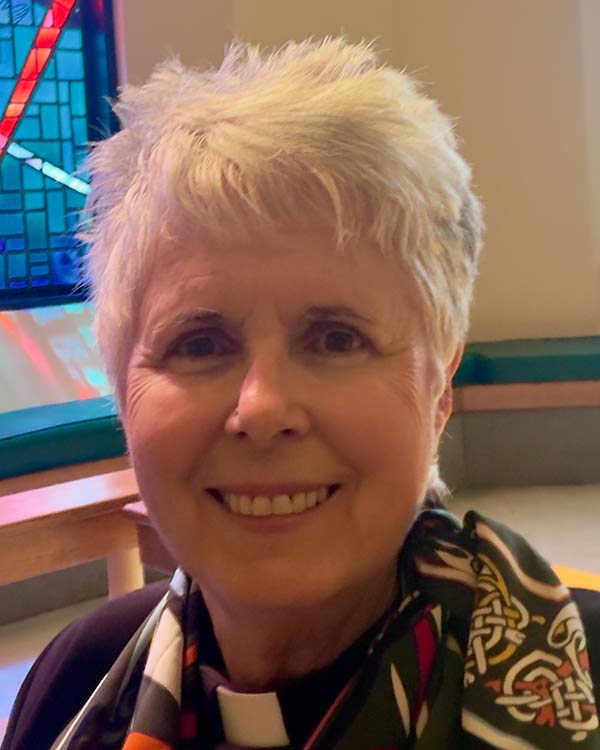 Portrait of a smiling clergy member with short white hair wearing a clerical collar and patterned scarf, seated near a stained glass window inside a church