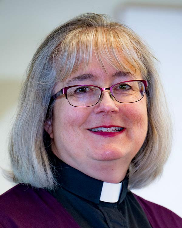 Portrait of a smiling clergy member with shoulder-length gray hair and glasses wearing a clerical collar and purple vestment against a light background