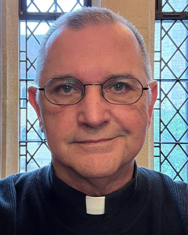 Portrait of a clergy member with short gray hair and glasses wearing a clerical collar, seated in front of leaded church windows