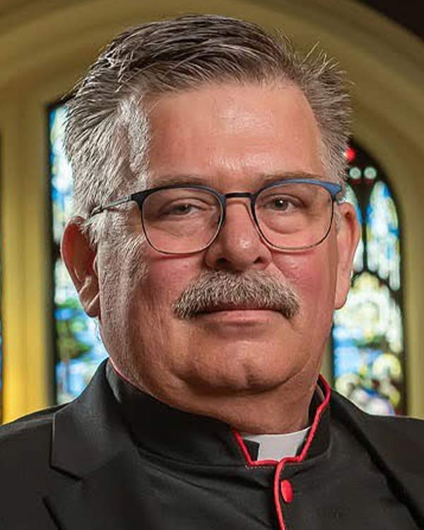 Portrait of a clergy member with gray hair, glasses, and a moustache wearing a clerical collar and black cassock, with stained glass windows in the background