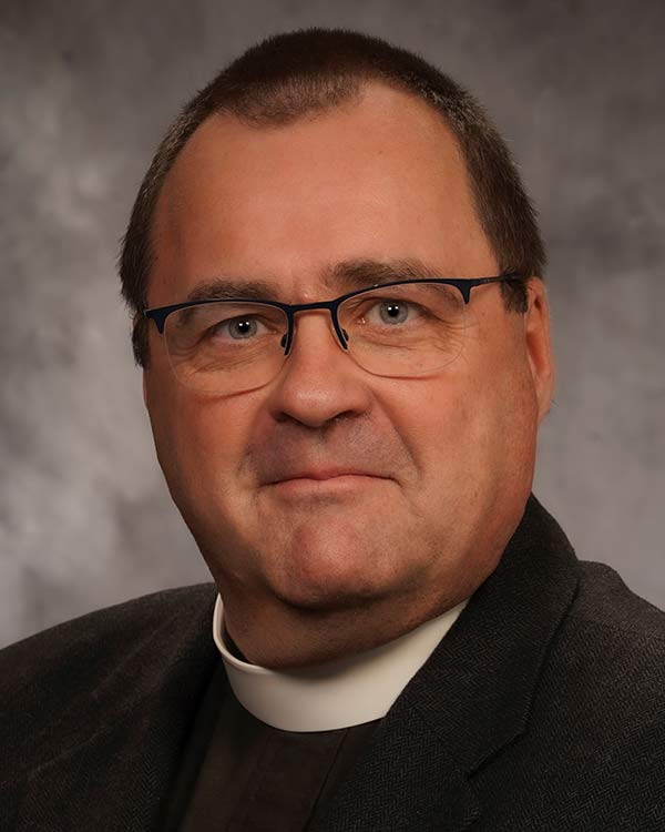Portrait of a clergy member with short dark hair and glasses wearing a clerical collar and dark jacket against a neutral studio background