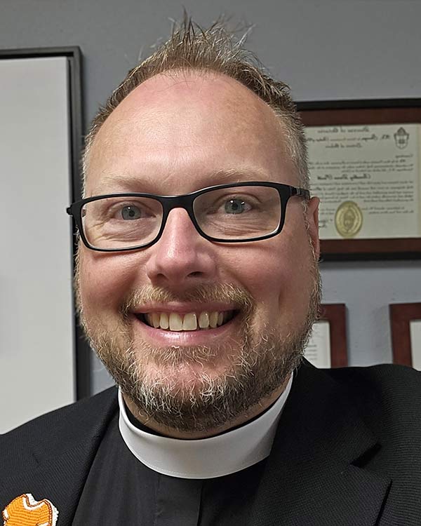 Portrait of a smiling clergy member with glasses and a beard wearing a clerical collar and dark jacket, with framed certificates on the wall behind him